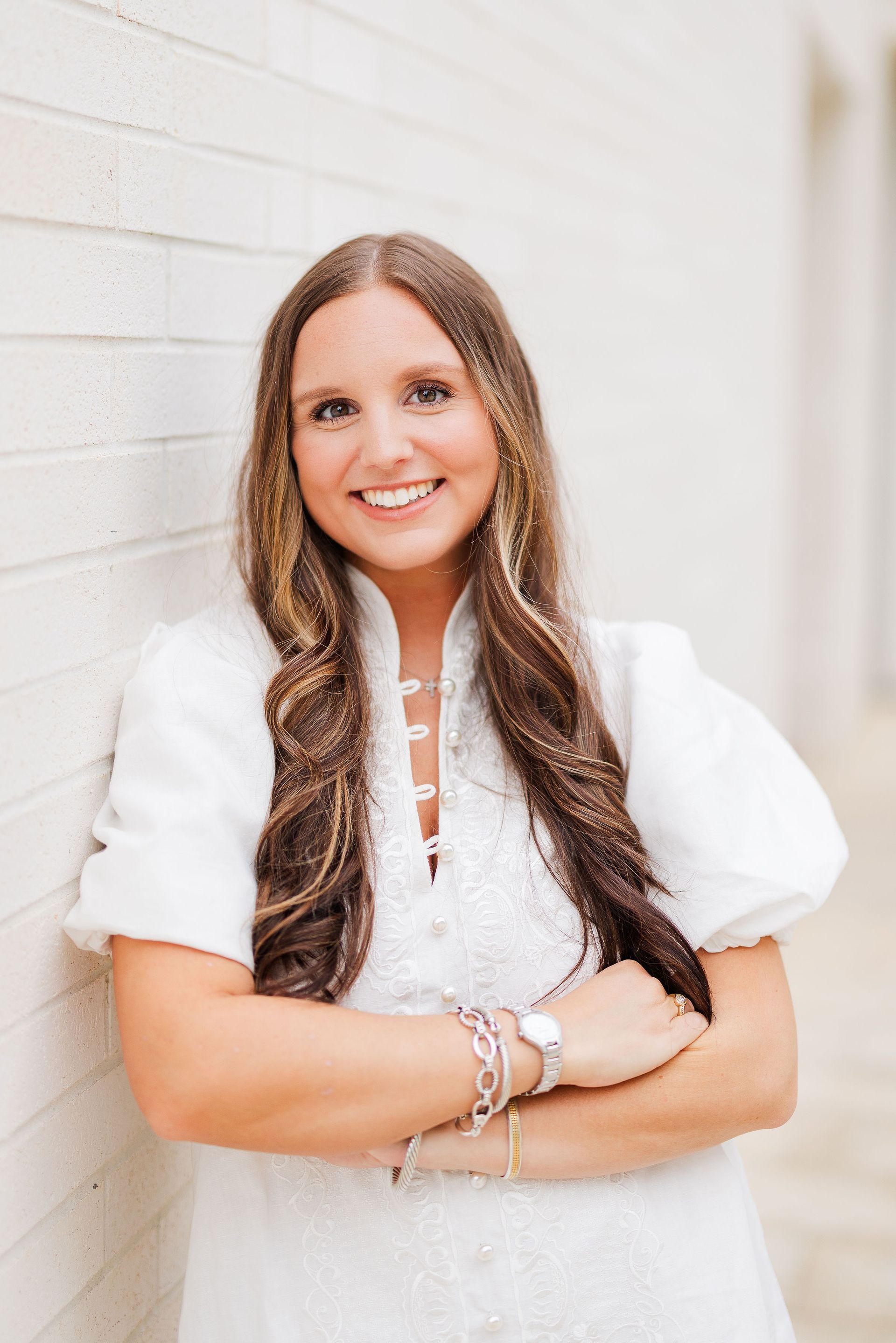 Smiling person in a white dress with long wavy hair, standing with arms crossed against a light wall