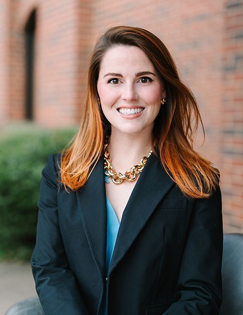 Smiling professional in a dark blazer and gold necklace, standing outdoors against a brick wall.