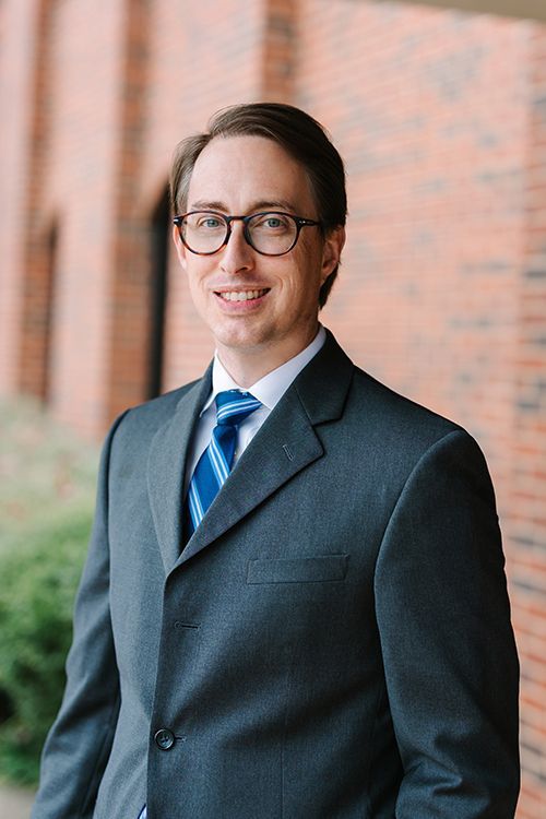 Man in a dark suit and blue tie standing outdoors by a brick wall, smiling at the camera