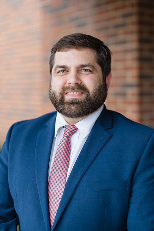 Portrait of a smiling man in a blue suit and red tie against a brick wall.