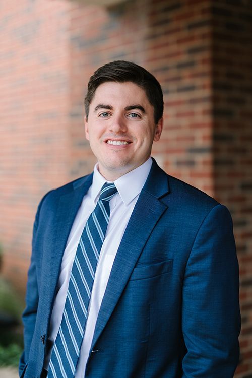 Man in a blue suit and striped tie smiling in front of a brick wall