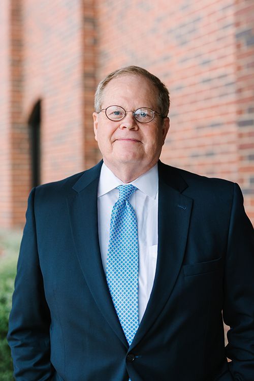 Man in a navy suit and blue tie standing outdoors against a brick wall, facing the camera.