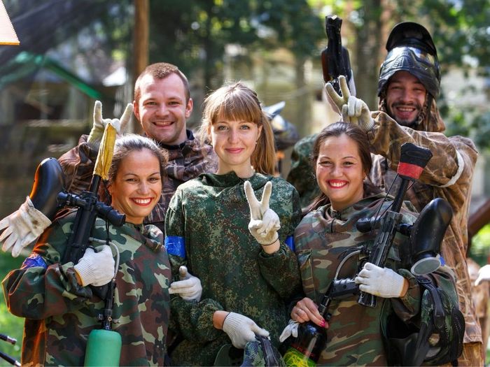 Groupe d'amis en tenue de camouflage souriant après une partie de paintball, équipés de lanceurs et masques de protection.