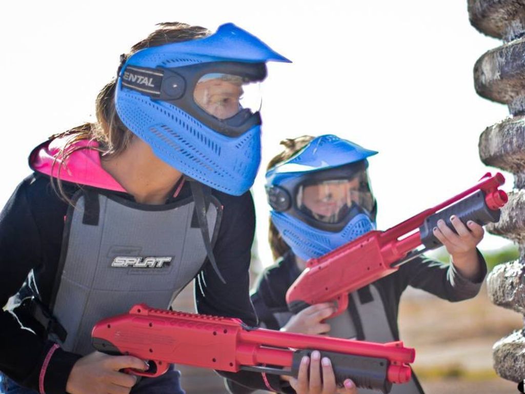 Deux enfants équipés de masques bleus et de lanceurs rouges, concentrés derrière un obstacle lors d’une partie de paintball
