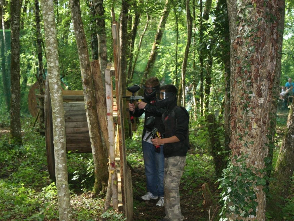 Groupe d’enfants en pleine partie de paintball, cachés derrière une barricade en bois, évoluant sur un terrain forestier.