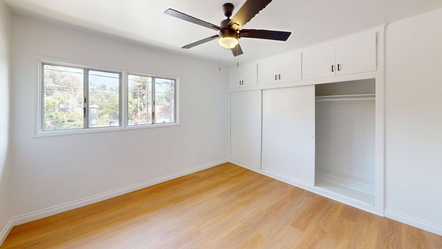 Bright empty bedroom with white walls, wood floor, ceiling fan, windows, and built-in closet cabinets