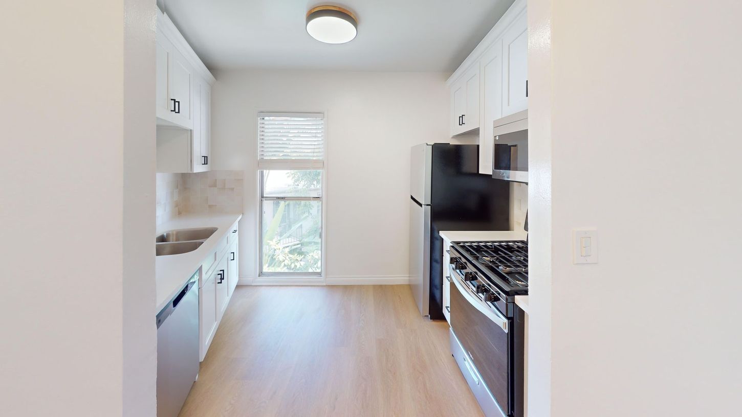 Bright narrow kitchen with white cabinets, light wood floor, window, and stainless steel stove and refrigerator
