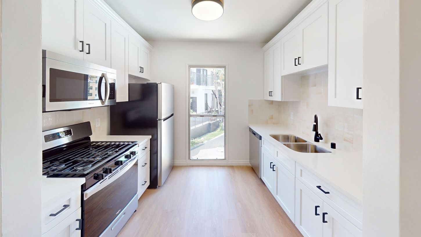 Bright narrow kitchen with white cabinets, stainless appliances, light wood floor, and glass door at the end.