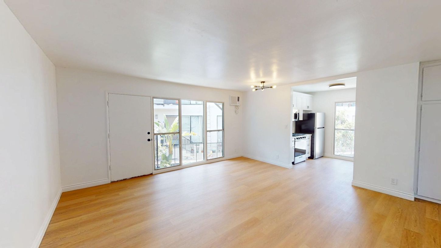 Bright empty living room with wood floors, white walls, balcony doors, and kitchen area.