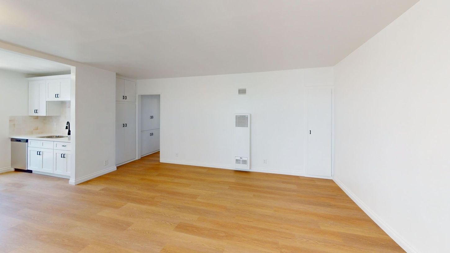 Empty white living room with light wood floors and open kitchen area on the left
