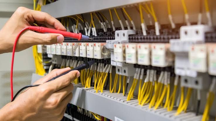 Hands testing circuit breakers in an electrical panel with yellow wires and a red multimeter probe