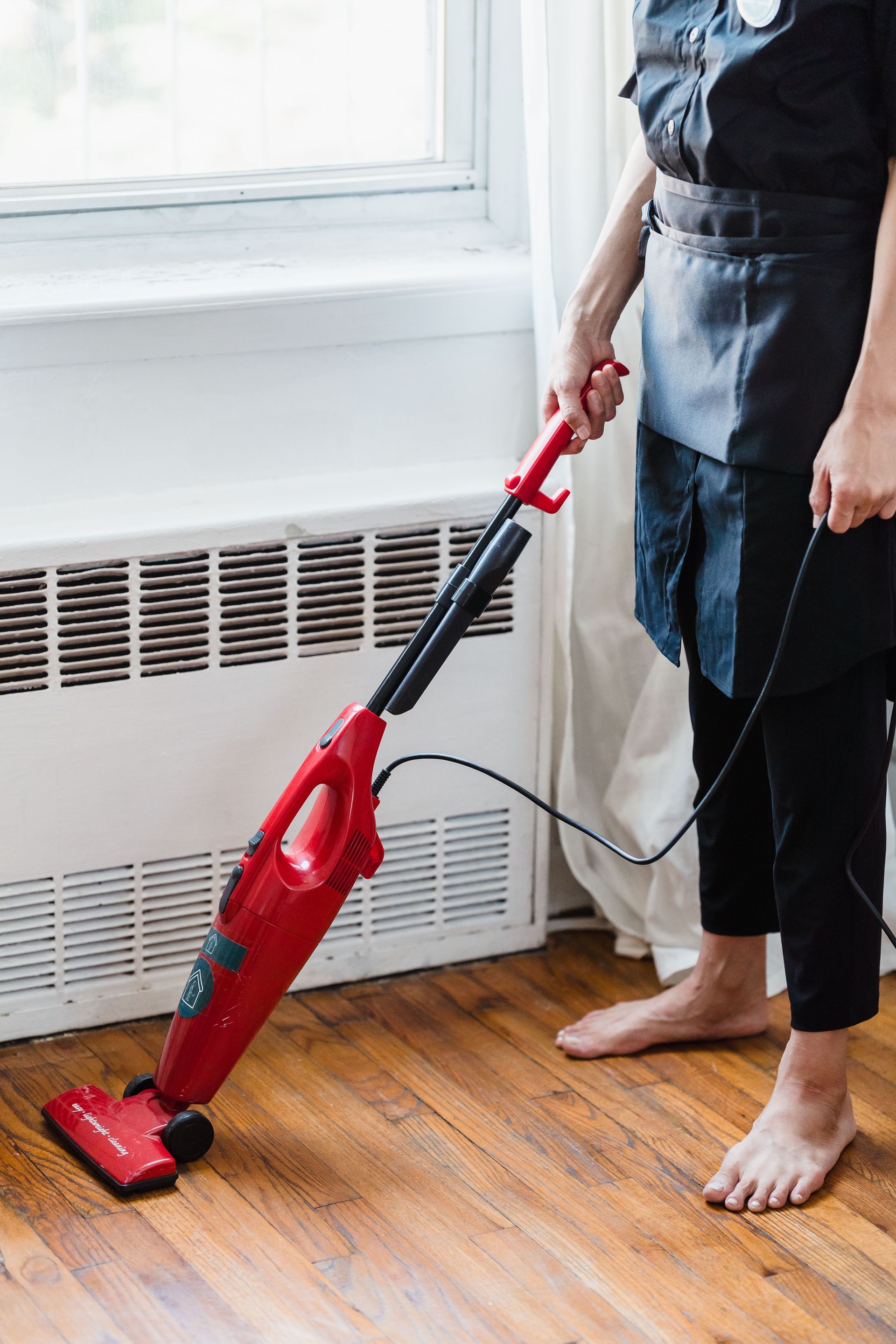 Person using a red cordless vacuum on a hardwood floor near a window and radiator