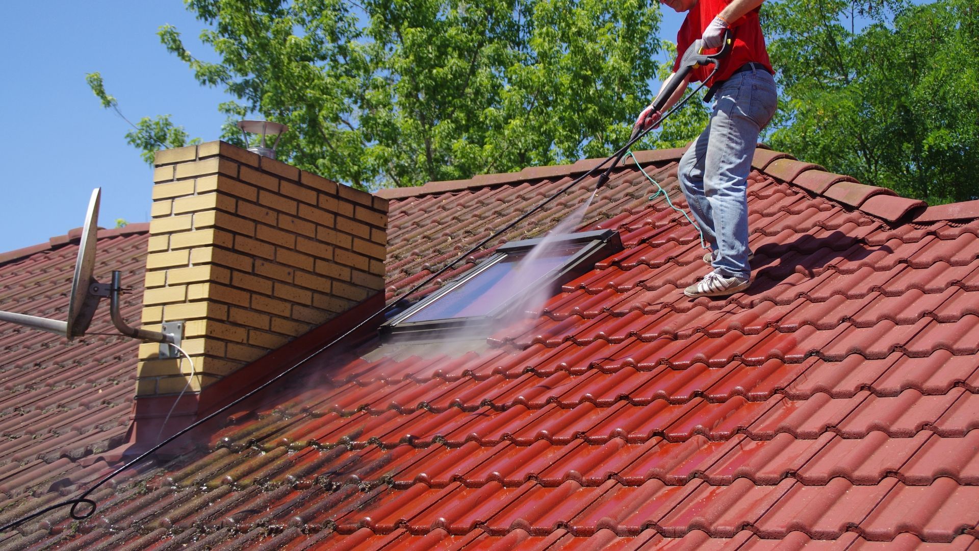 Worker pressure-washing a red-tiled roof beside a brick chimney on a sunny day