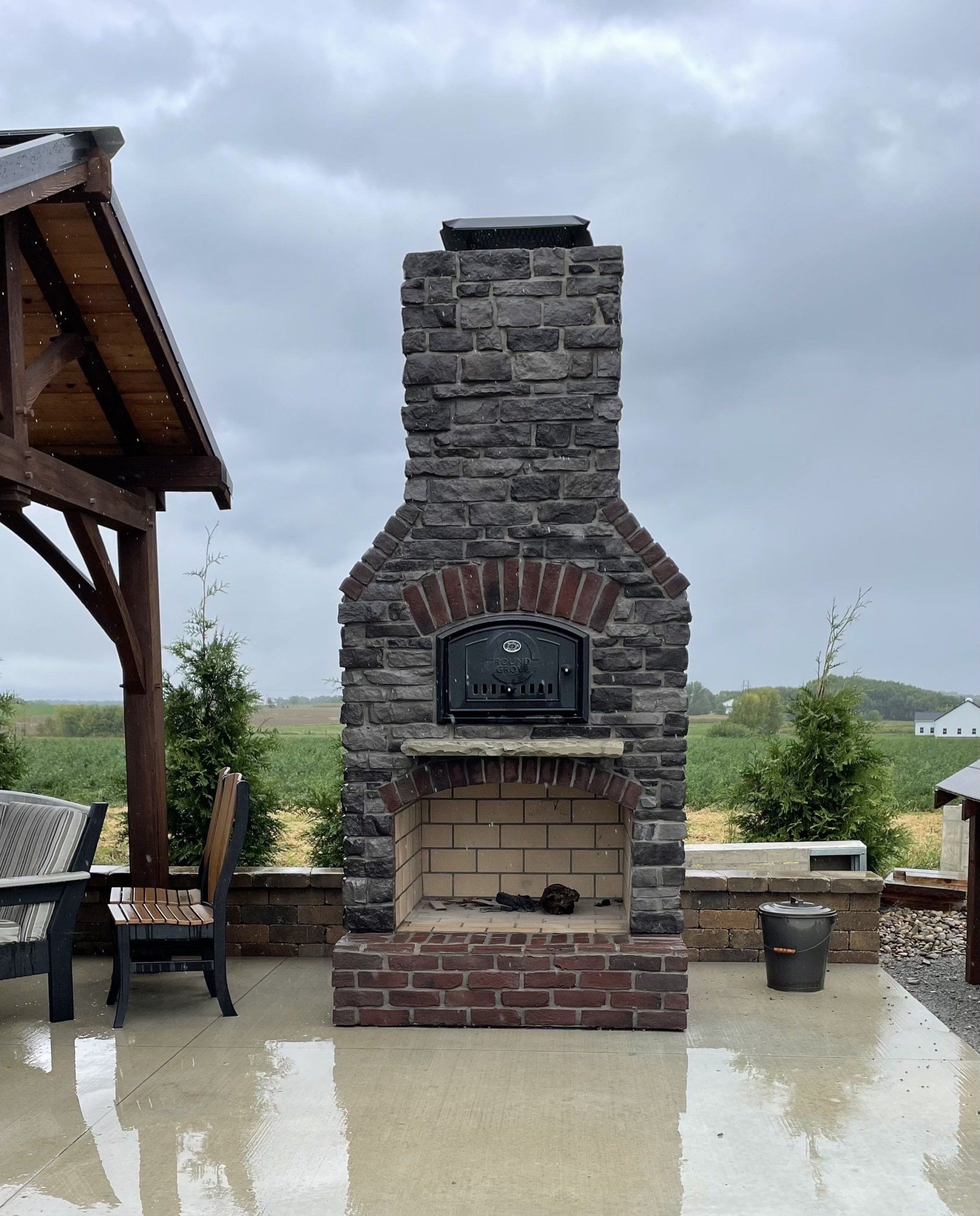 Outdoor brick pizza oven with a dark grey stone chimney and red brick accents on a rainy patio.