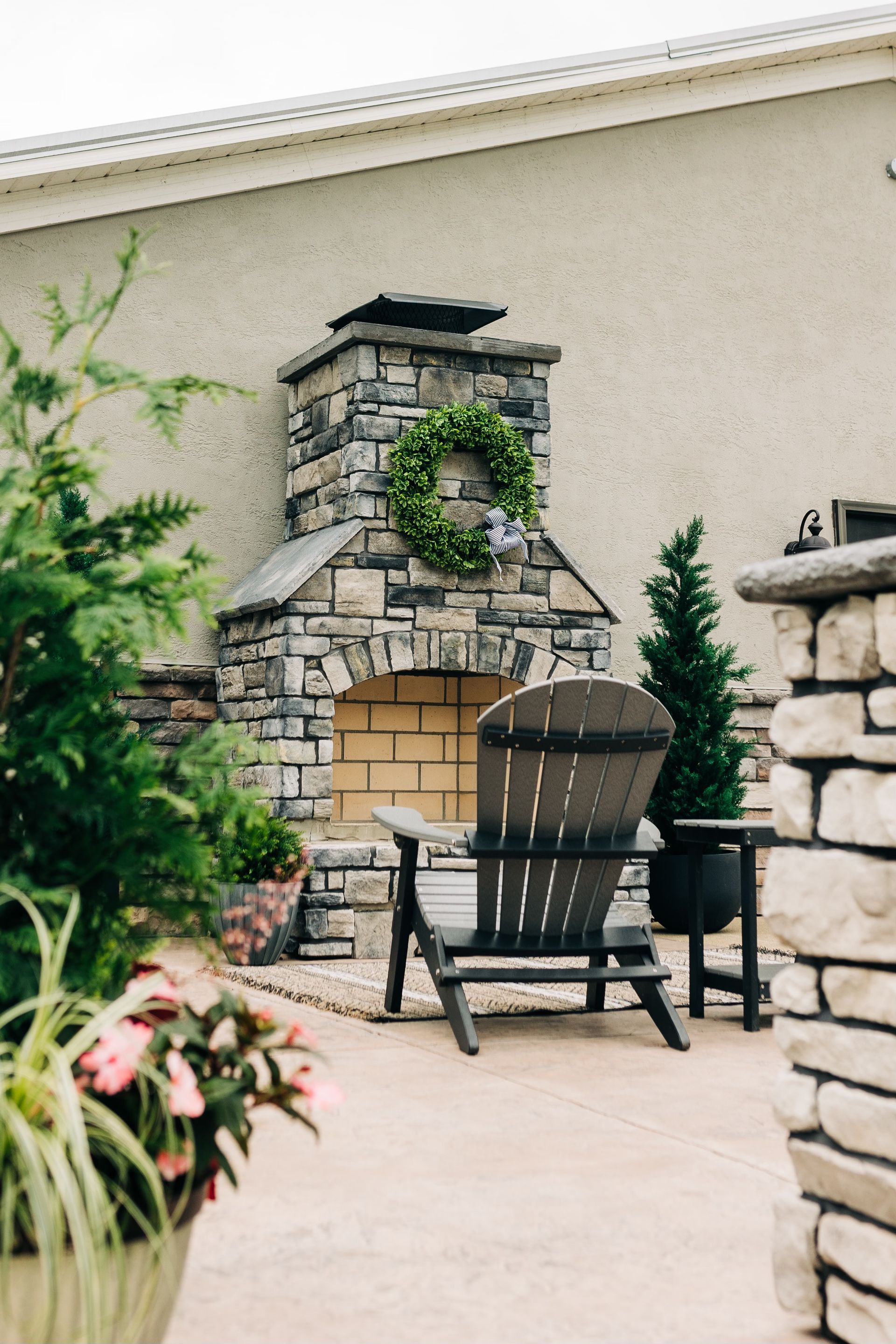 Stone fireplace with a green wreath, Adirondack chair, and potted plants on a patio.