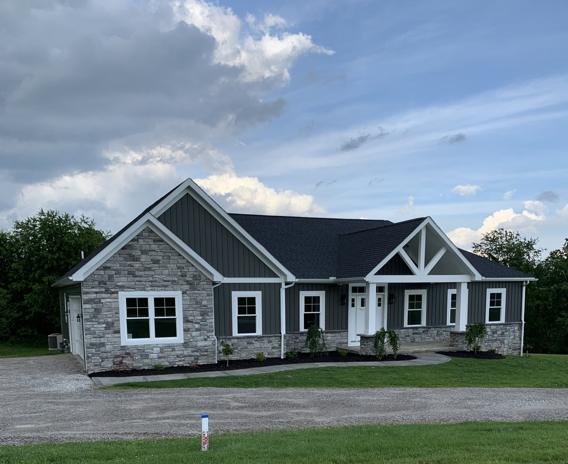 Gray and stone-faced ranch-style house with dark gray siding, white trim, and a black roof under a cloudy sky.