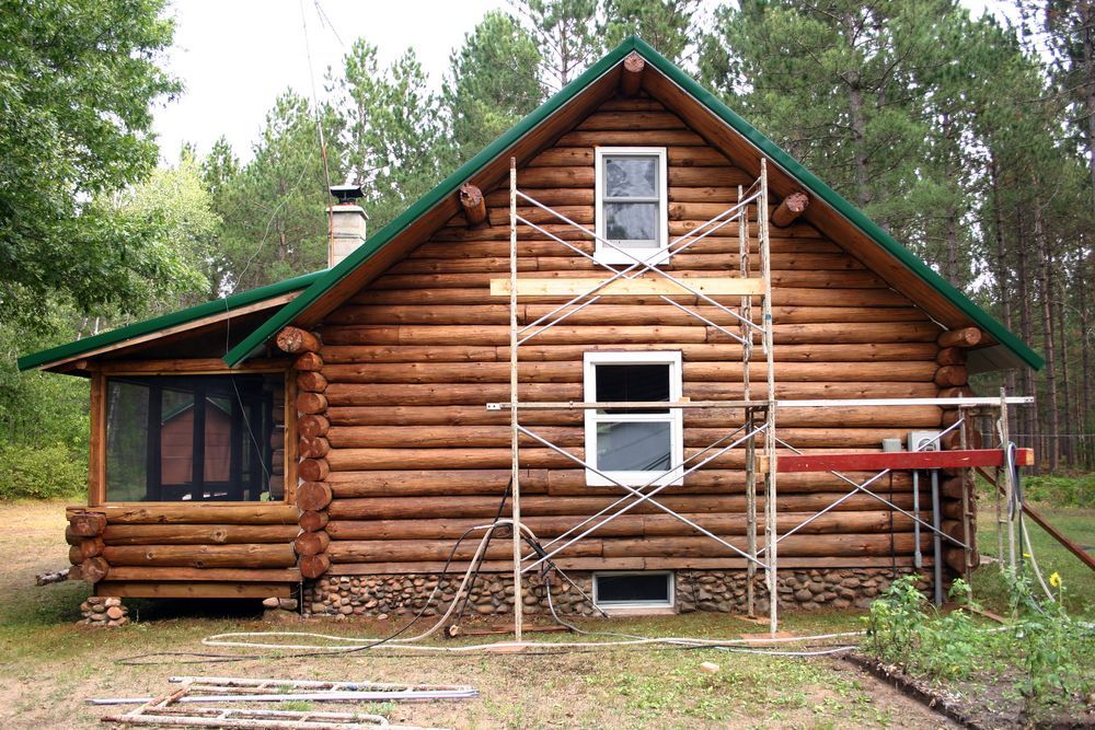 Log cabin with green roof trim and scaffolding on the front, surrounded by trees and grass