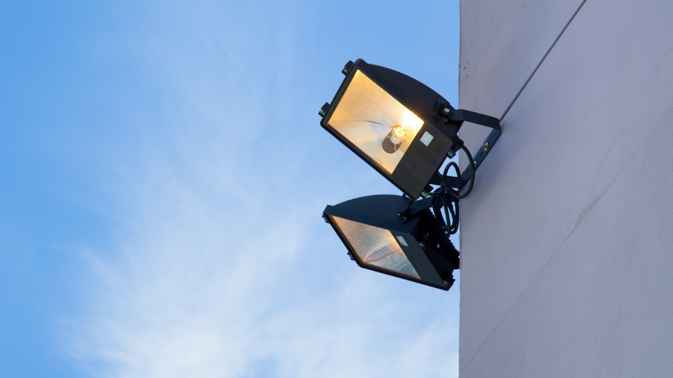 Two bright floodlights mounted on a light-colored wall against a blue sky.