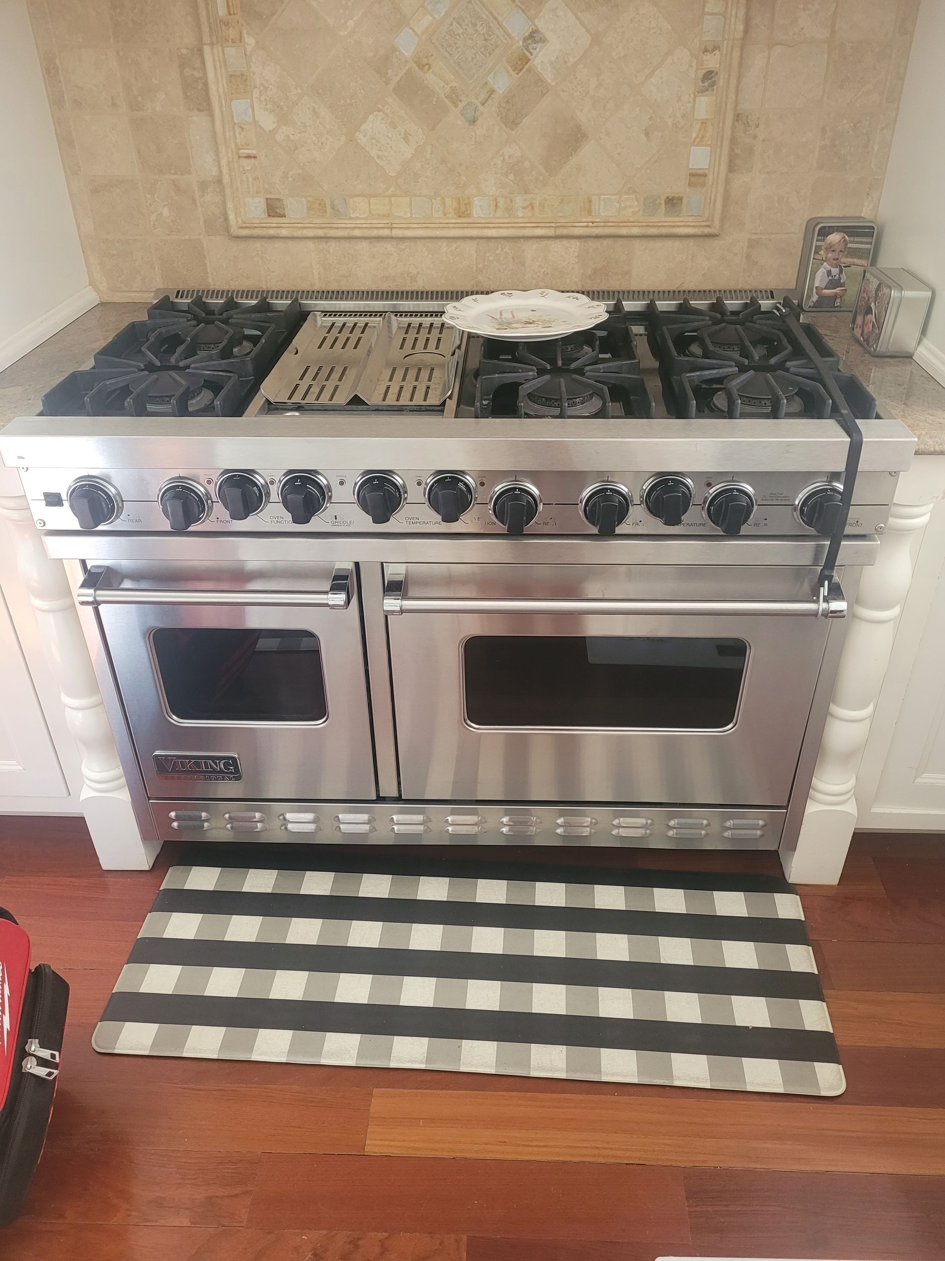 A person lies on a wooden floor, reaching under a stainless steel dishwasher to adjust the kickplate.