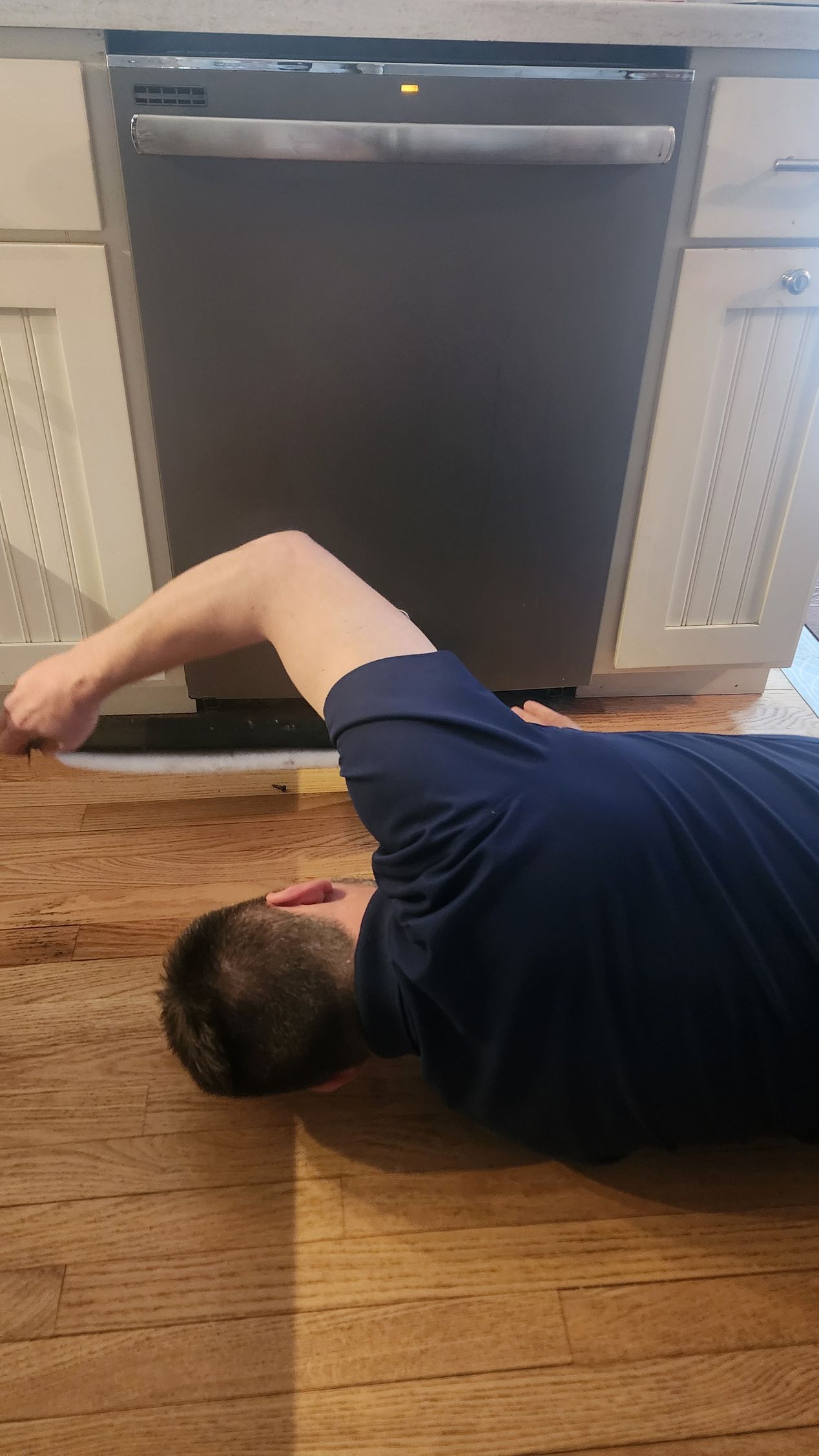 A person lies on a wooden floor, reaching under a stainless steel dishwasher to adjust the kickplate.