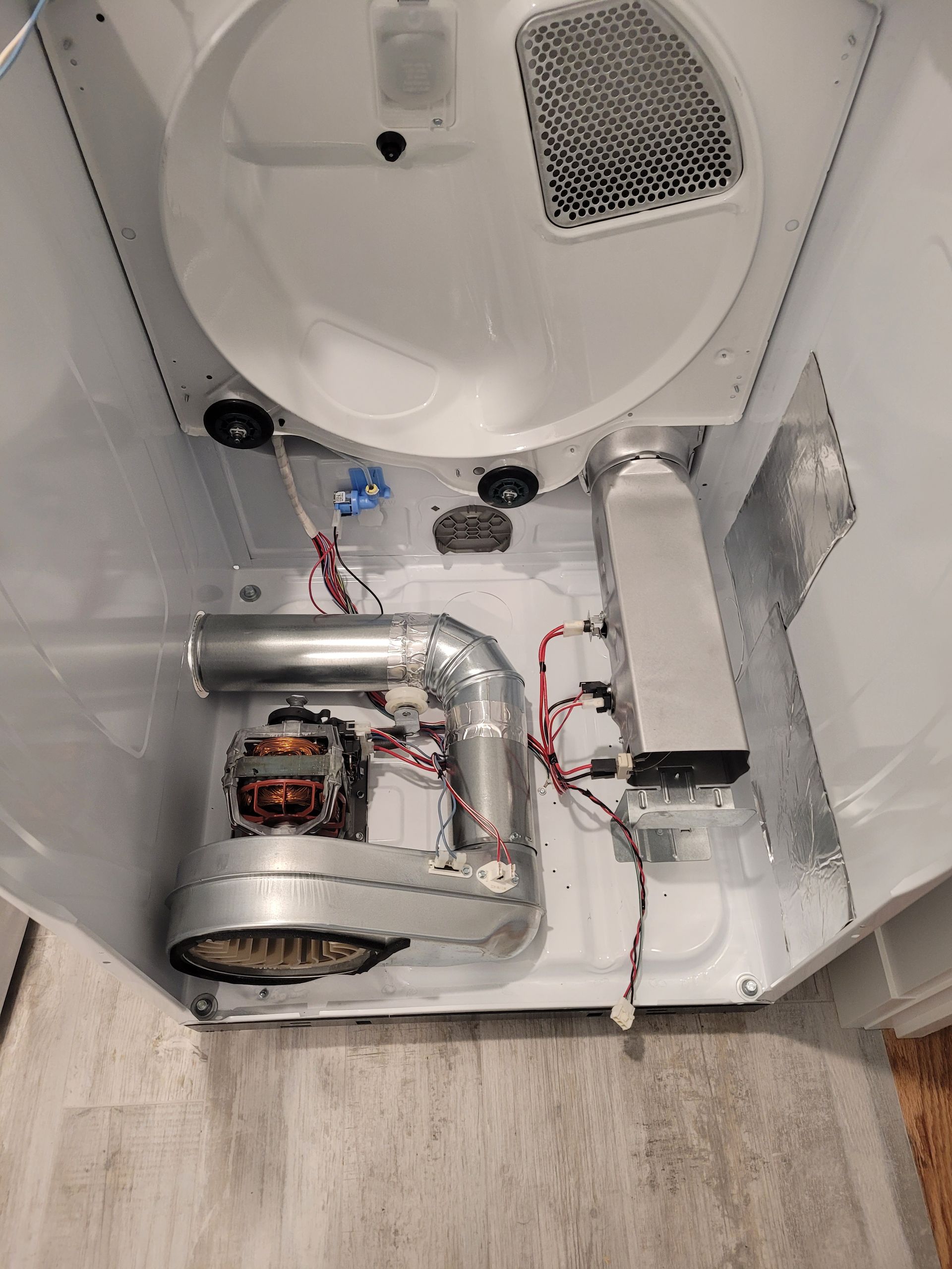 A person lies on a wooden floor, reaching under a stainless steel dishwasher to adjust the kickplate.