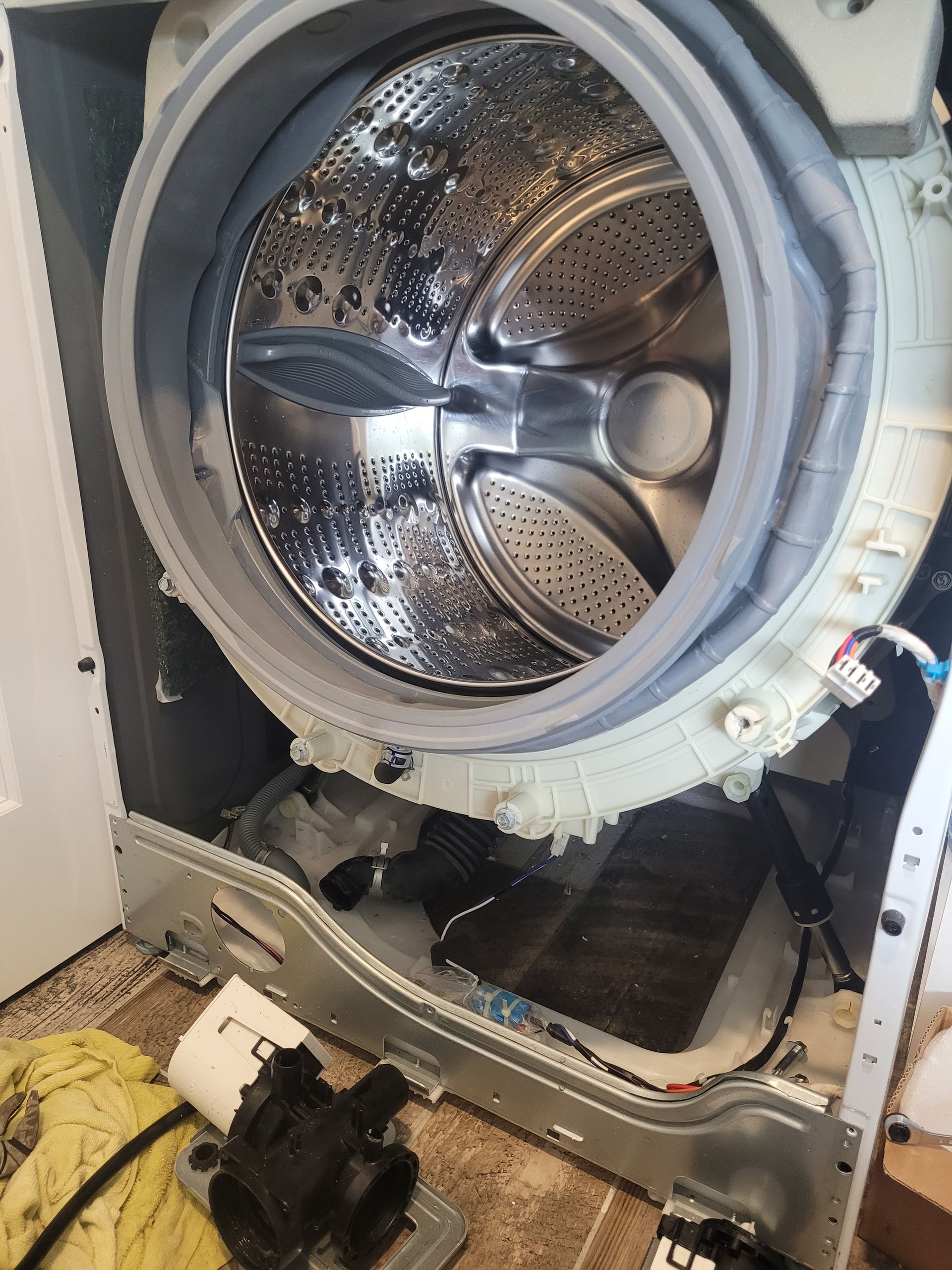 A person lies on a wooden floor, reaching under a stainless steel dishwasher to adjust the kickplate.
