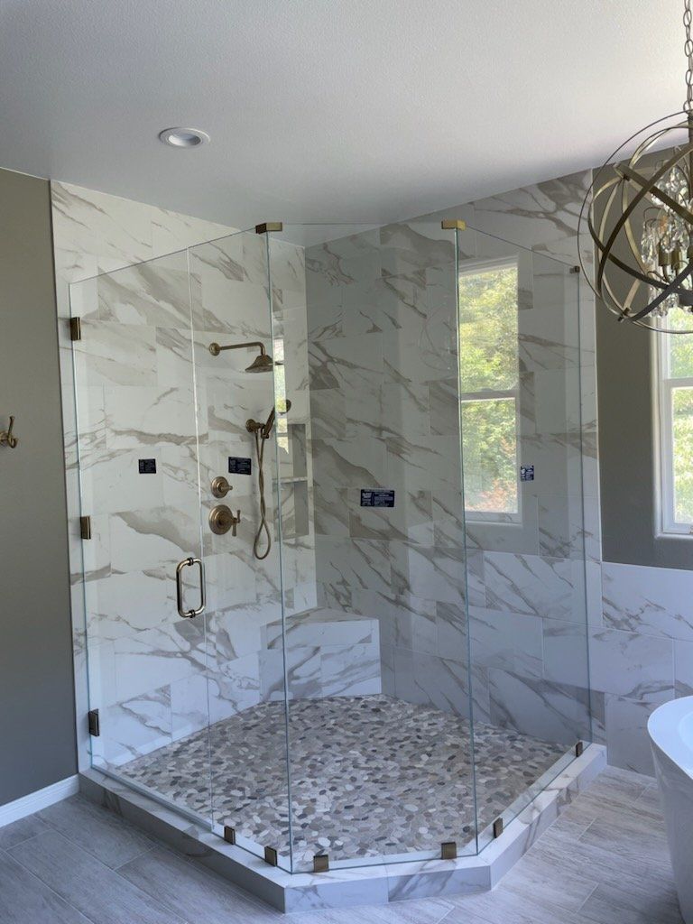 Glass-enclosed corner shower with white marble tile walls, pebble floor, gold fixtures, and a window.