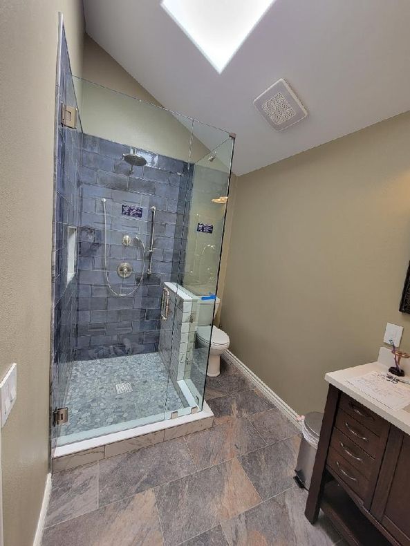Bathroom with glass shower, grey tile, toilet, vanity, and a skylight.