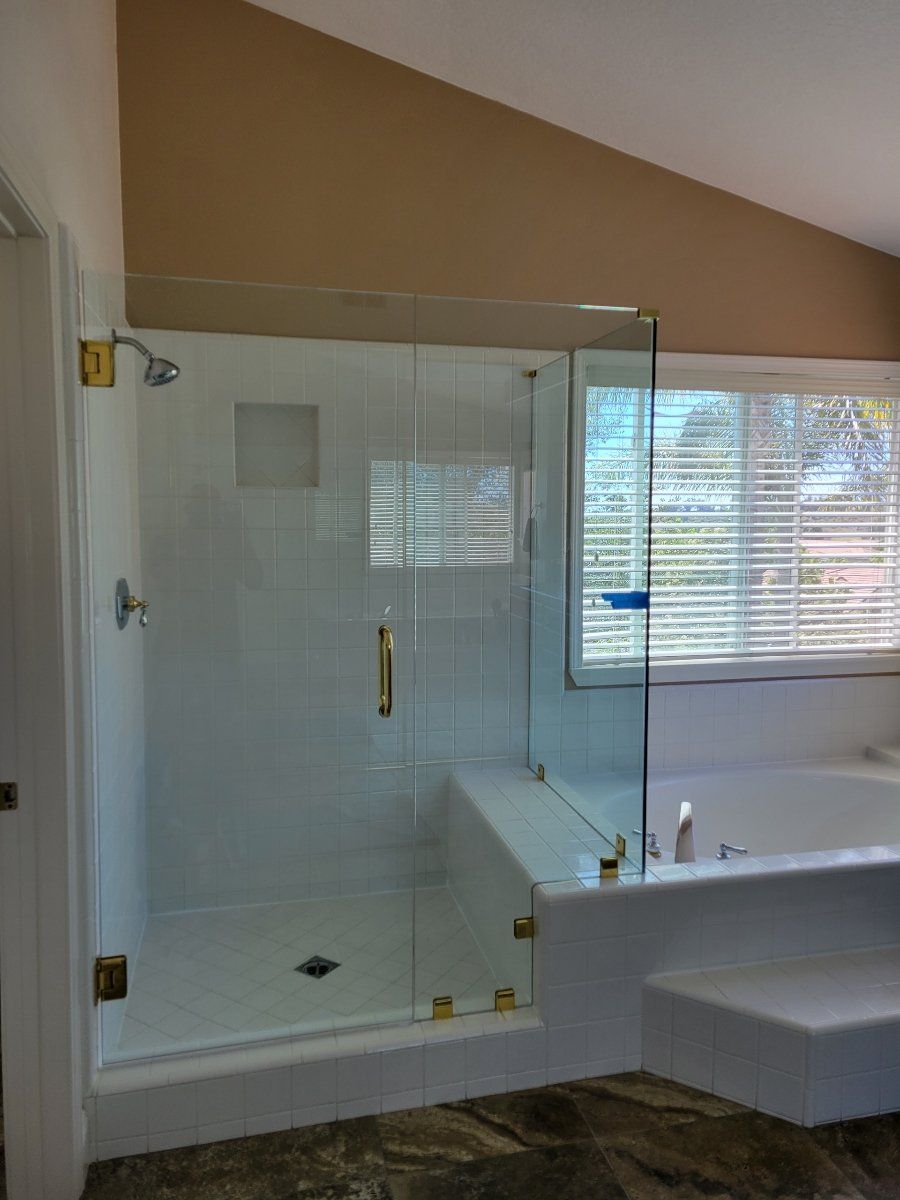 Glass shower enclosure with golden hardware next to a bathtub, white tile, and a window with blinds.