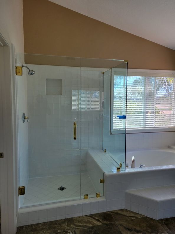 Glass shower enclosure with gold fixtures, white tiled walls, next to a bathtub with a window.