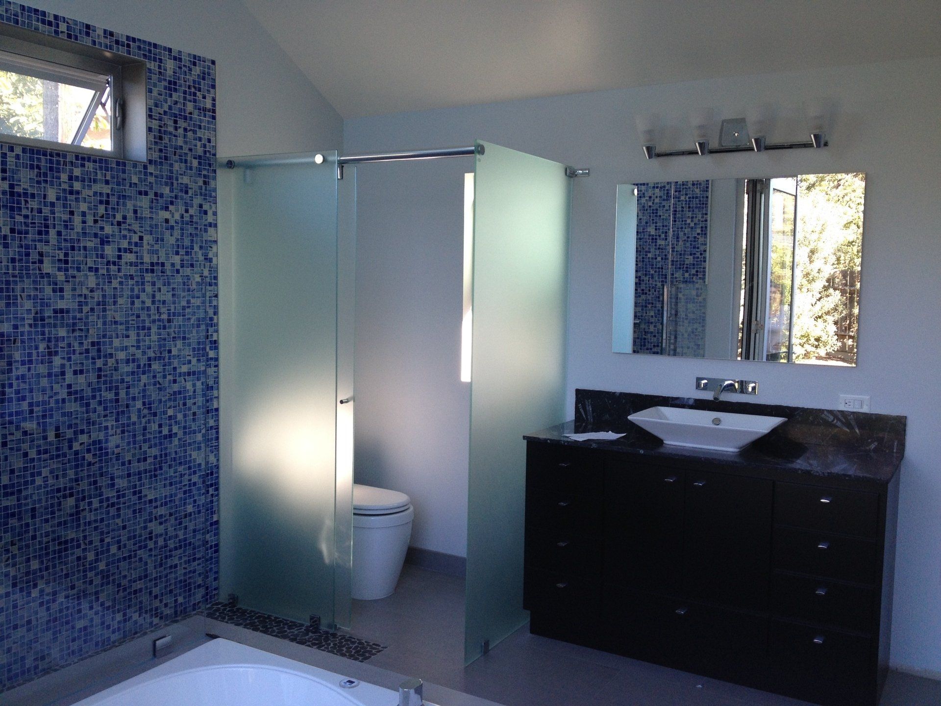 Modern bathroom with blue tile wall, frosted glass shower, dark vanity, and white sink.