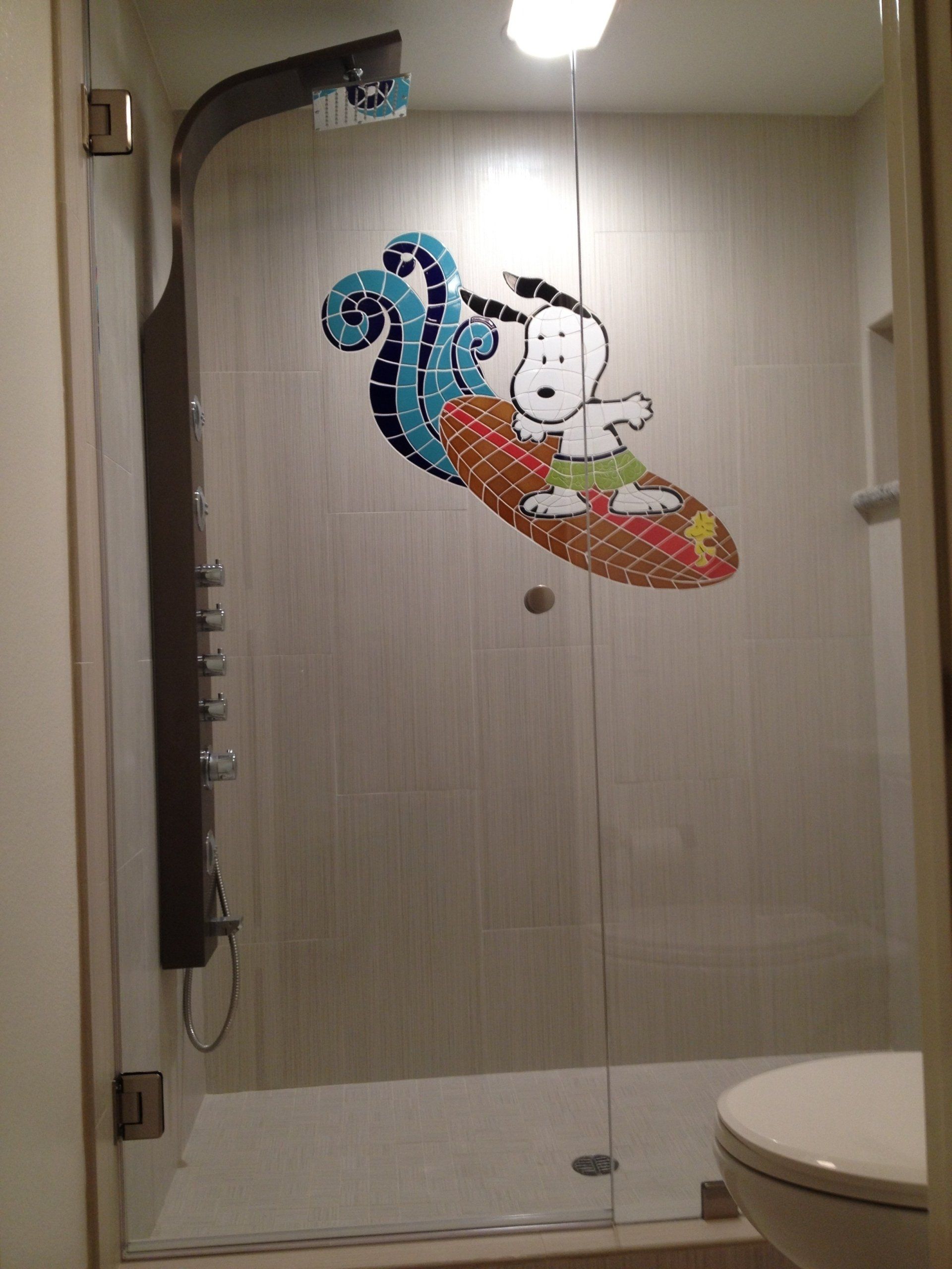 Shower with Snoopy surfing on the glass door, blue wave, tiled design, light gray walls, and a toilet.