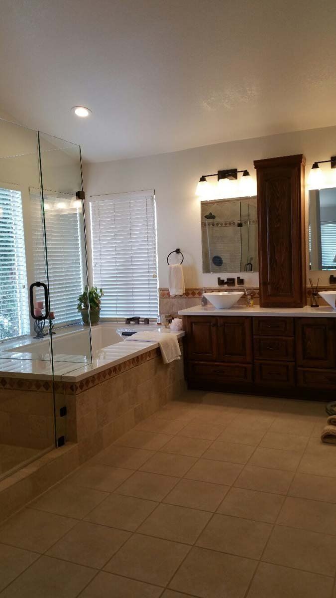 Spacious bathroom with tub, double sinks, wooden cabinets, and tile floor. Natural light streams through window blinds.