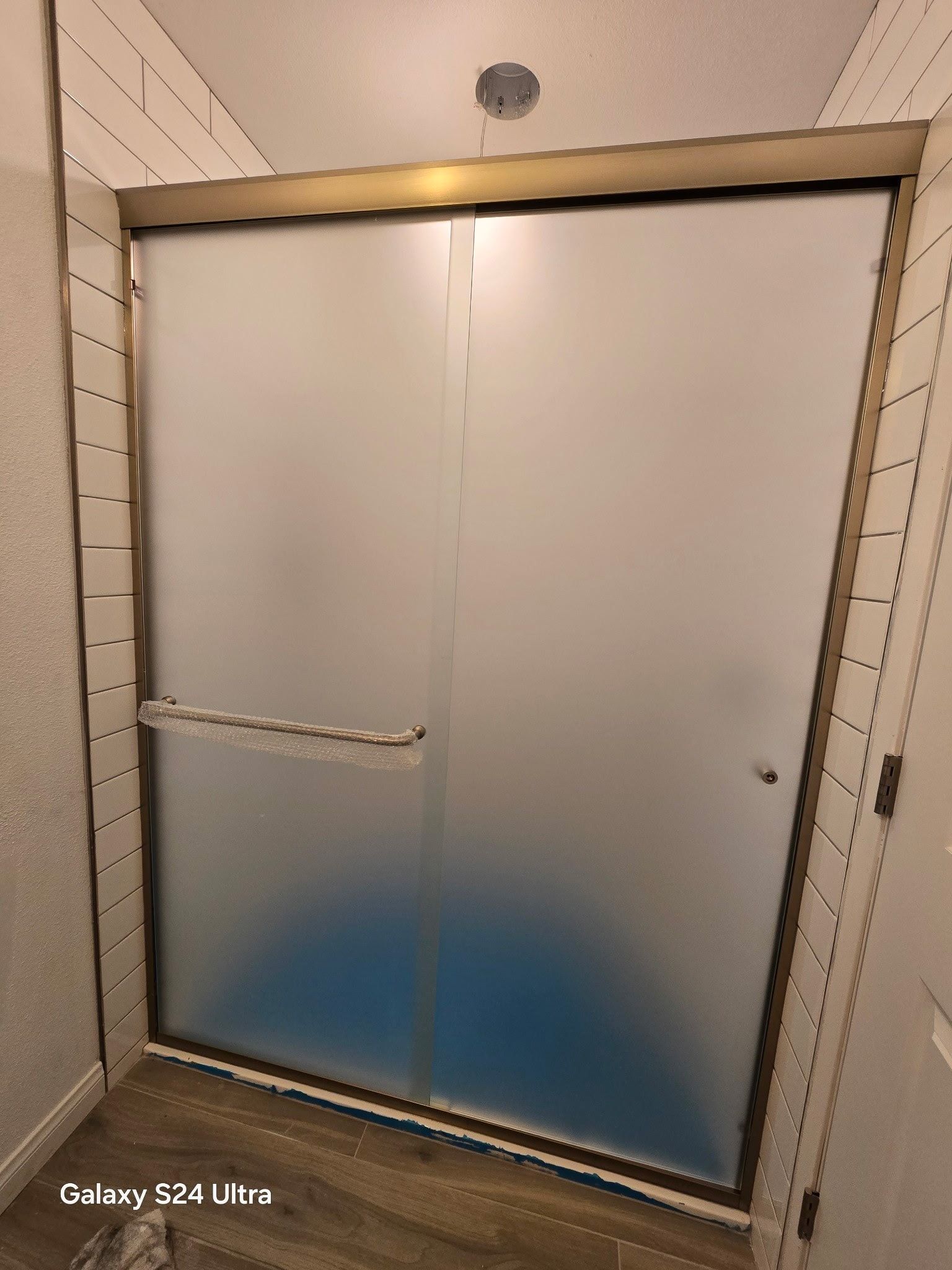 Frosted glass shower door with gold frame, white tiled walls, and wood-look flooring.