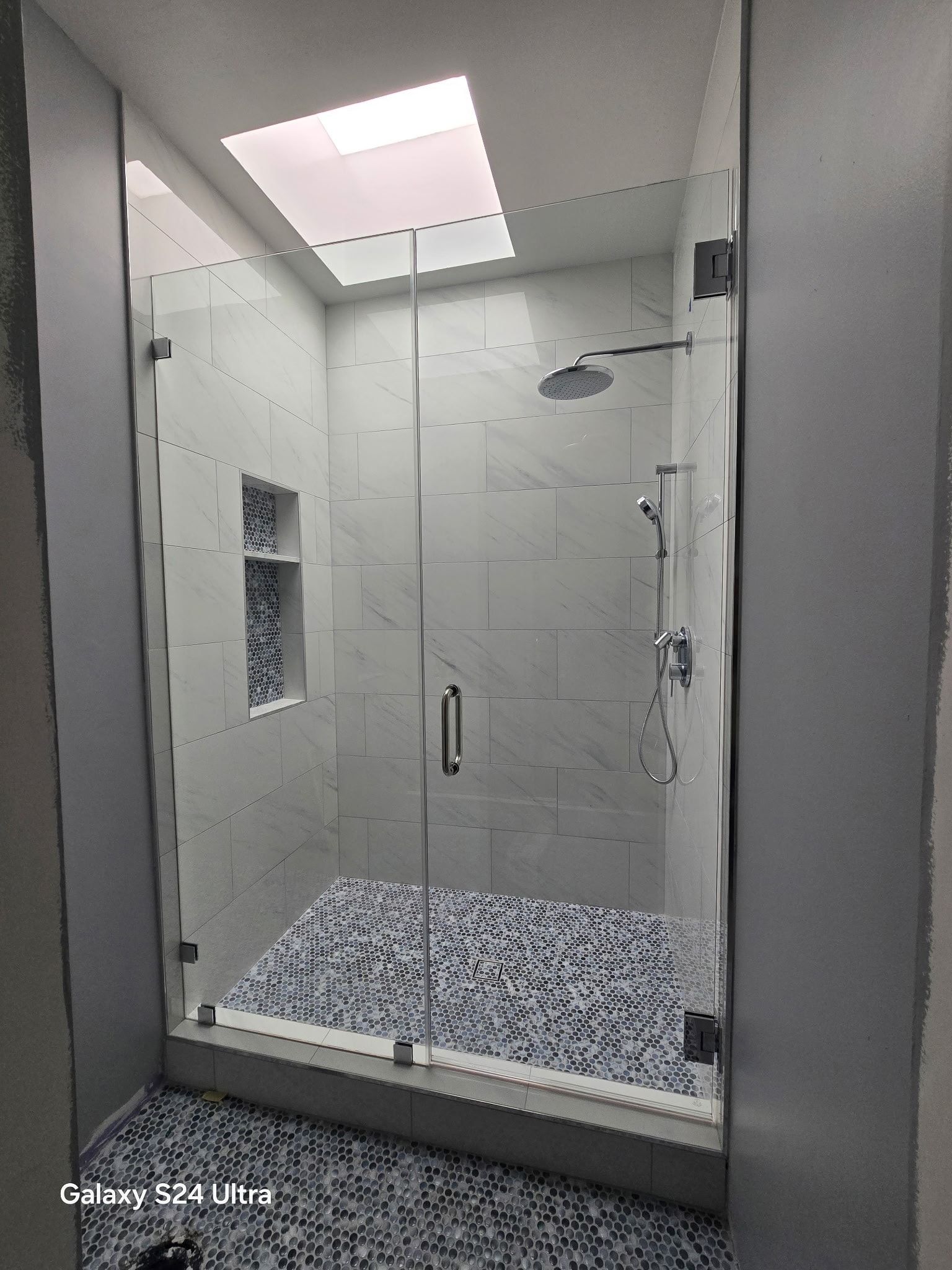 Glass shower with a tiled floor, white walls, and a skylight.