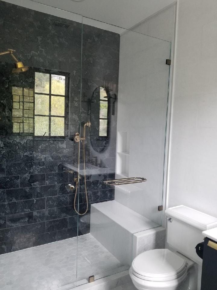 A modern bathroom with a glass shower, black tile wall, white bench, and toilet.