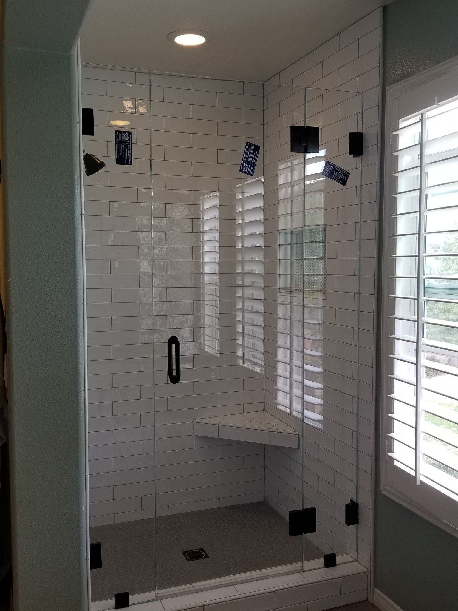 Glass shower with white tile, dark hardware, and a window with blinds.