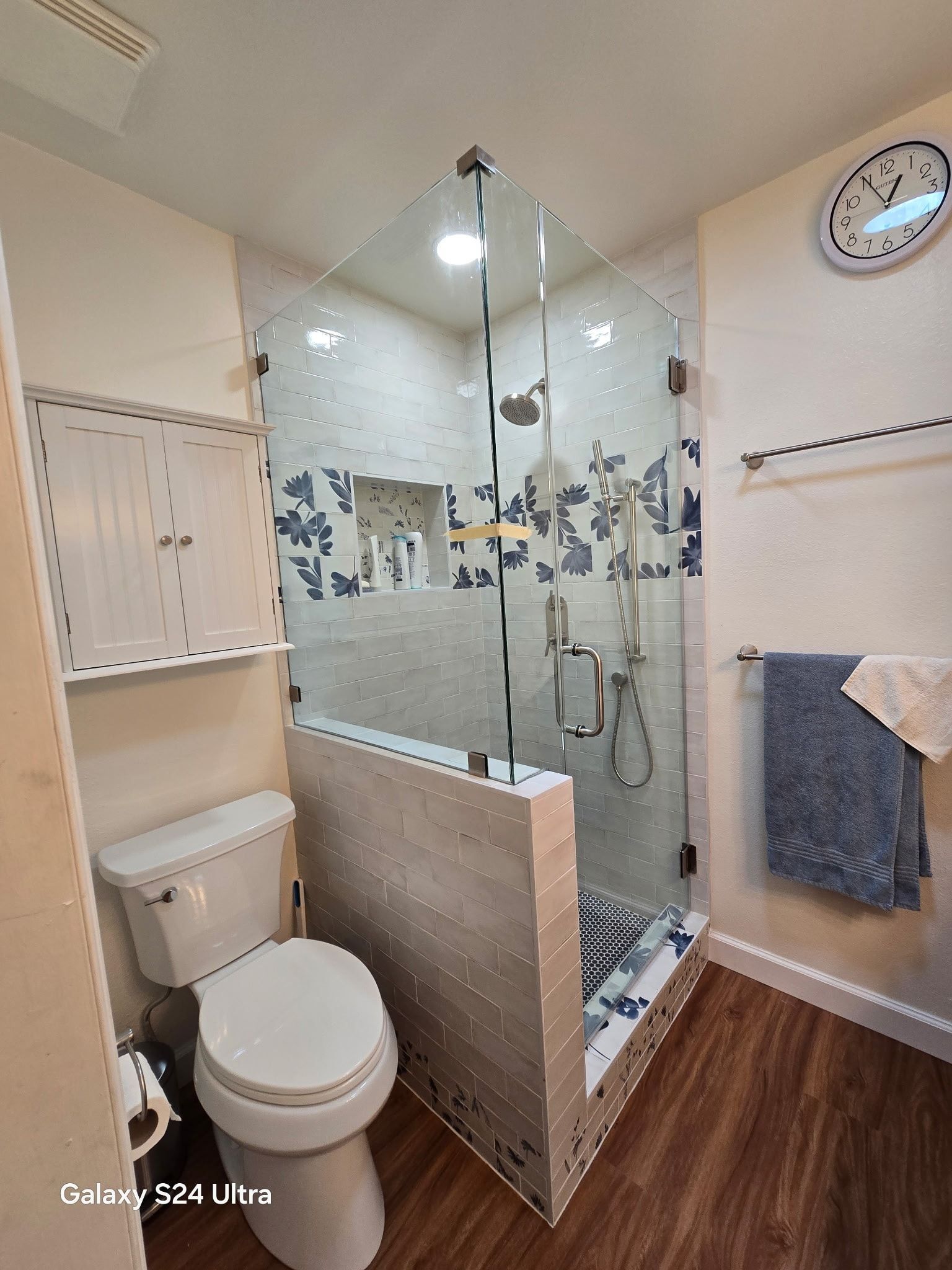 Bathroom with a glass shower, toilet, and wood-look floors; blue and white floral tiles.