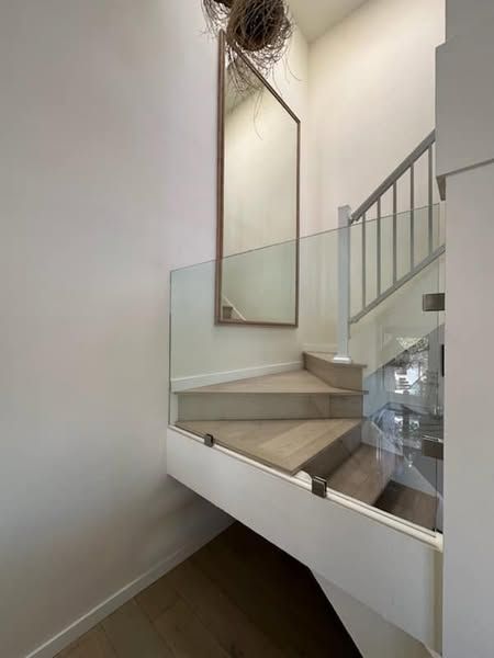 Staircase with wooden steps, glass railings, and a large mirror on the wall. Bright, airy interior.