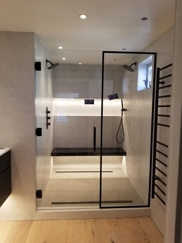 Modern shower with black fixtures, glass doors, and bench. Light wood floor.