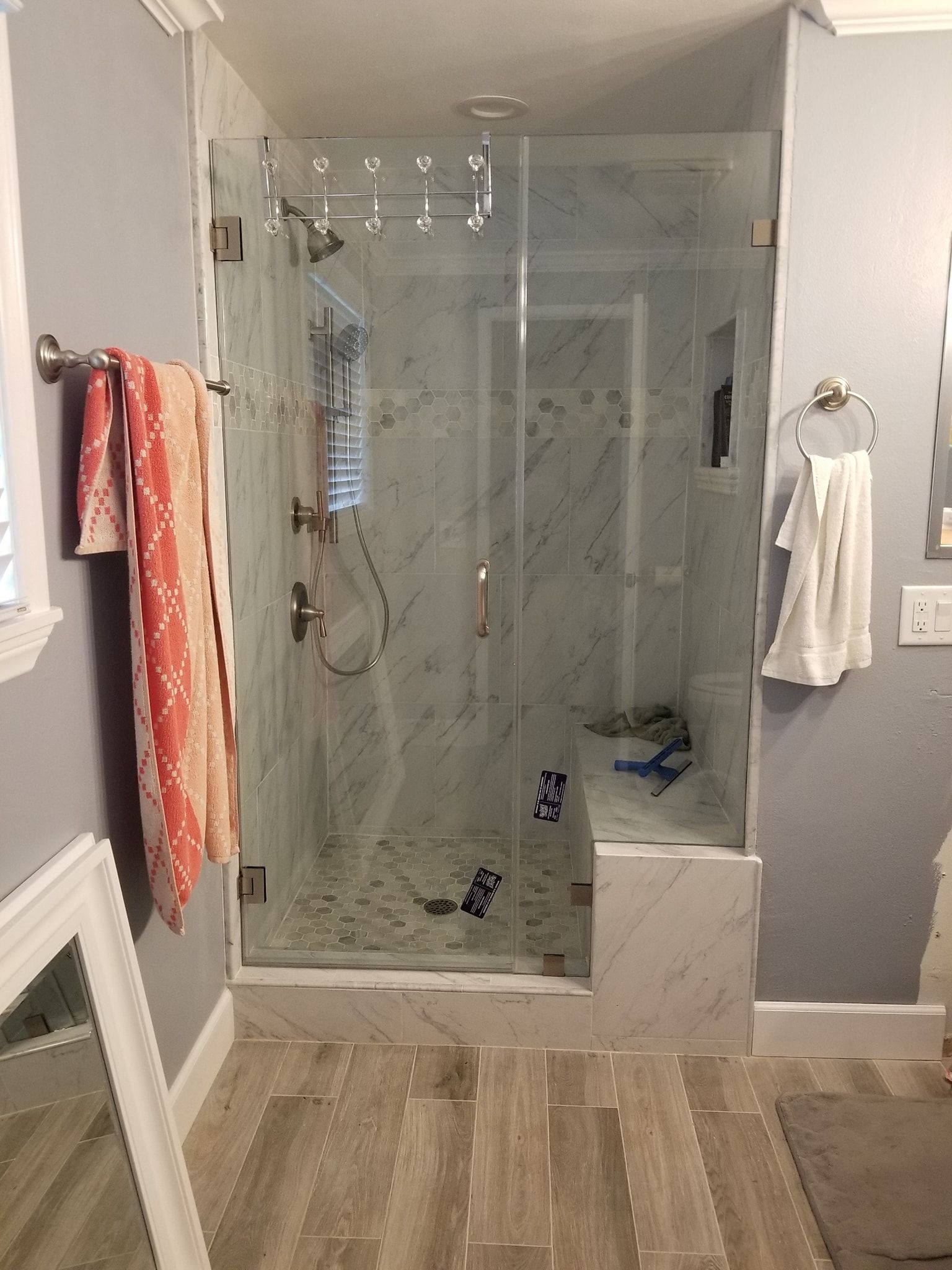 Bathroom with marble-tiled shower, glass door, and bench. Towels on a rack; light blue walls and floor tiles.