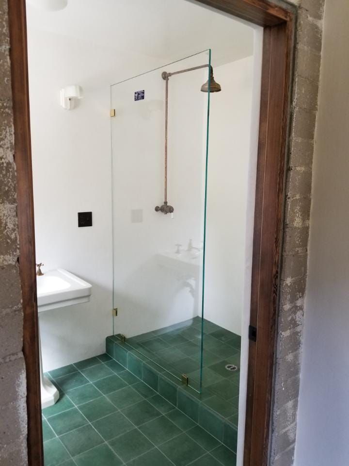 A shower with a glass door, green tile floor, white walls, and a brass showerhead.