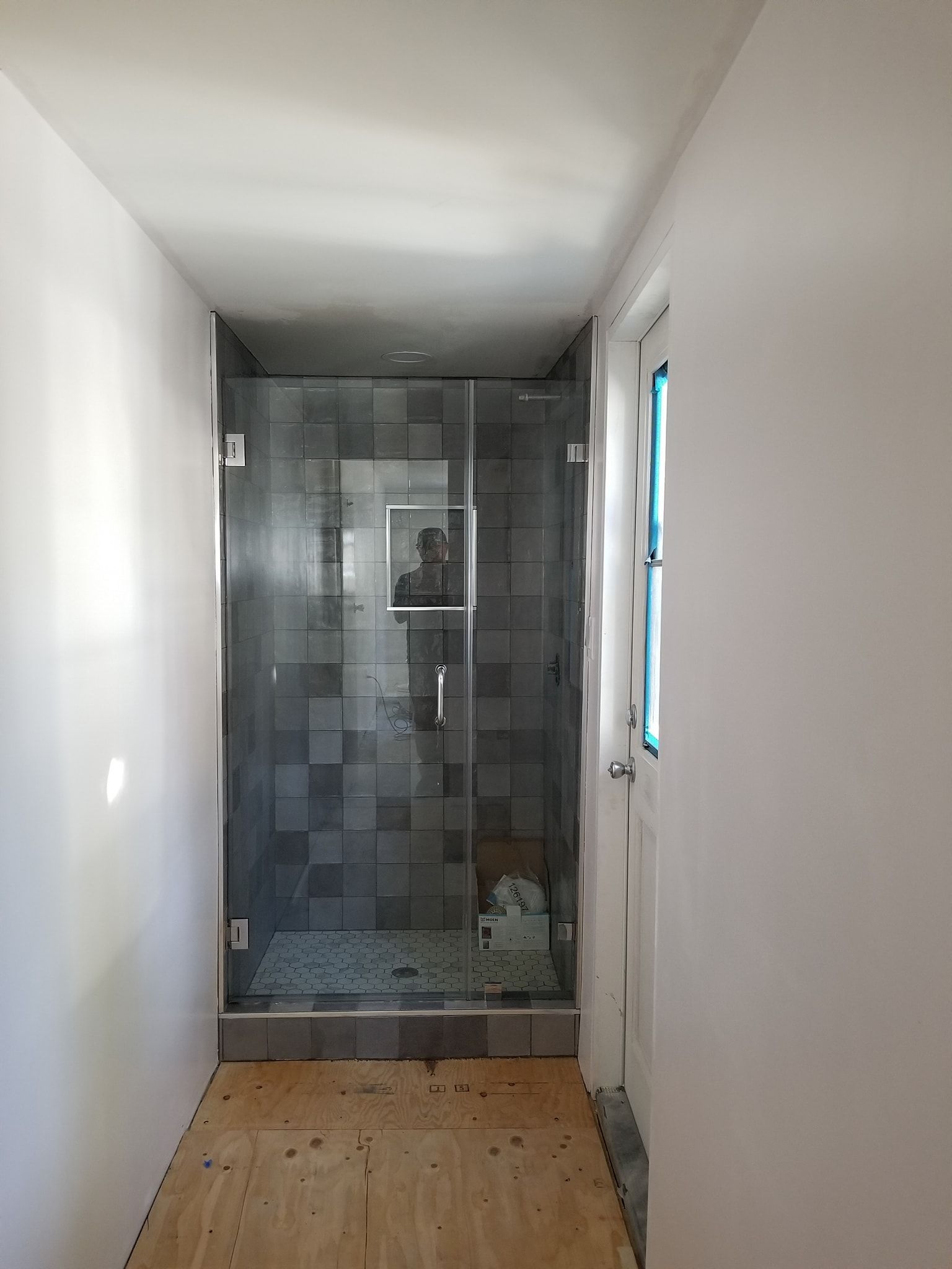 A narrow bathroom hallway with a glass shower enclosure. Dark tile inside the shower, unfinished floor.