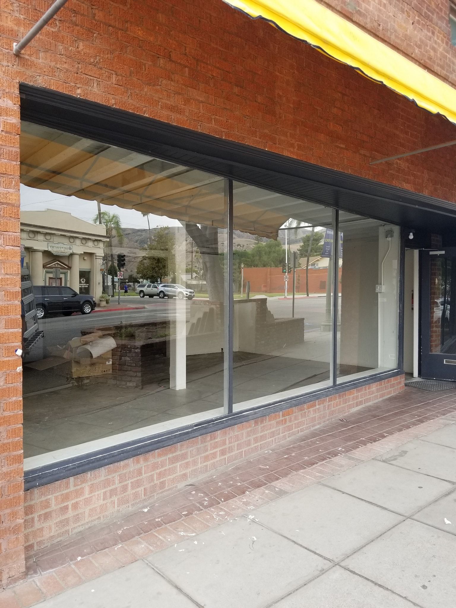 Empty storefront with large windows reflecting street scene. Brick building, yellow awning.