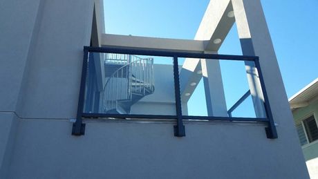 Glass balcony with black frame on a gray building, sunny day, spiral staircase in background.