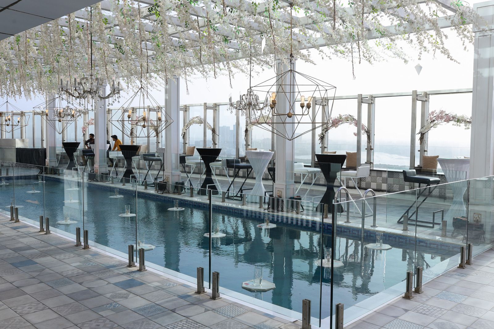 Rooftop pool with glass railing and decorative florals; tables and chairs set for a gathering with city views.