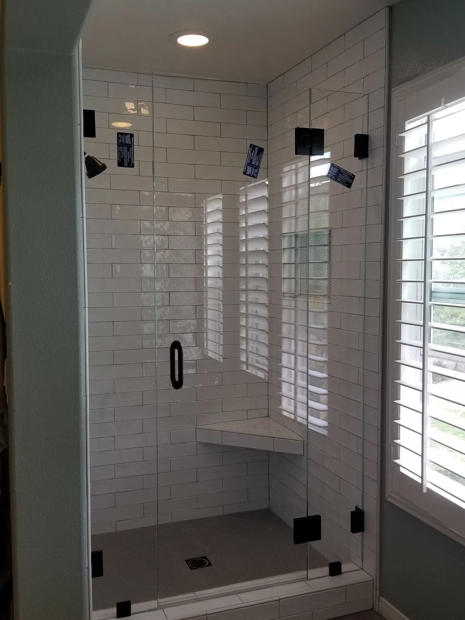 Glass shower enclosure with white tile, built-in bench, and bright window blinds