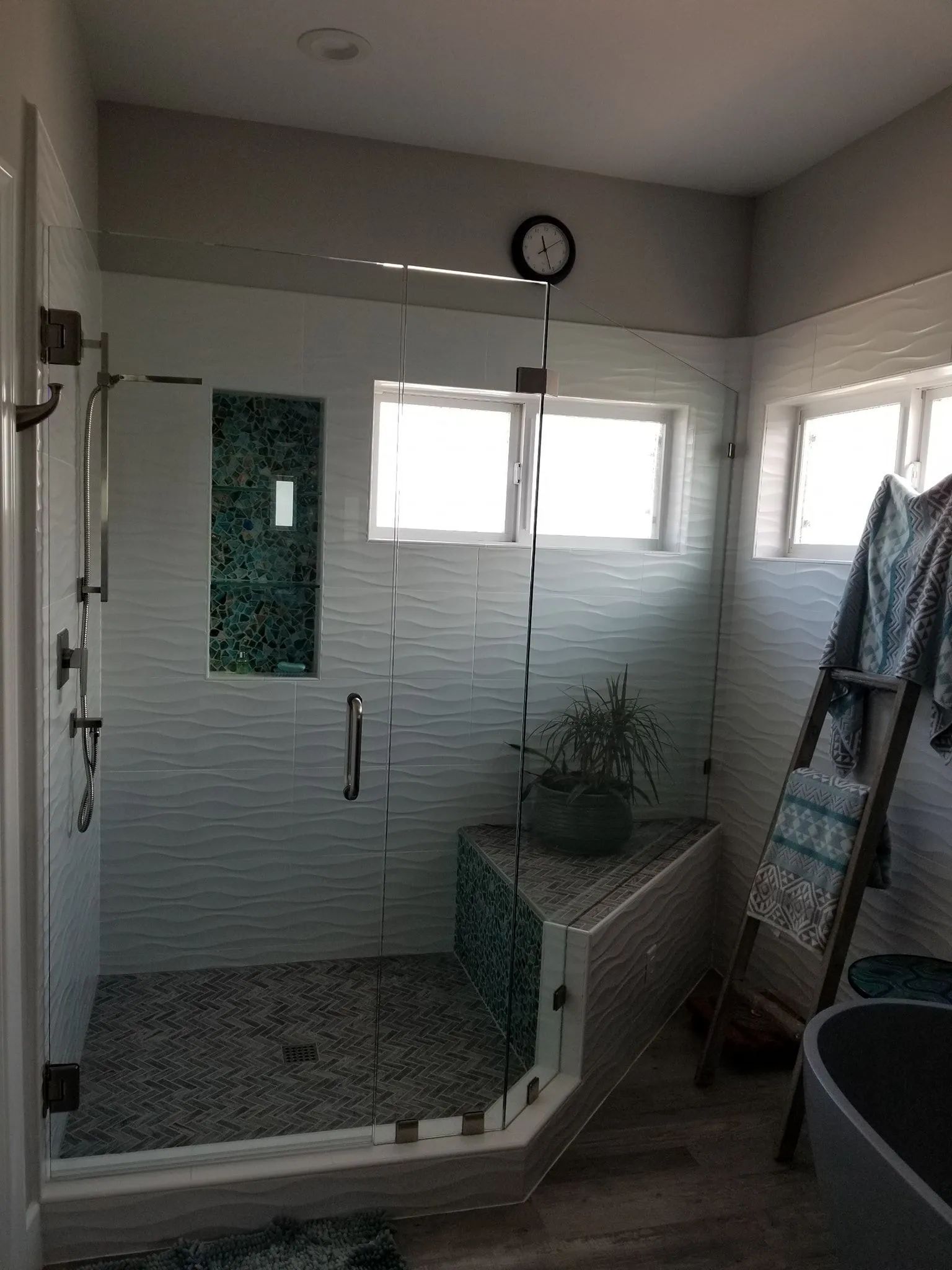 Bathroom with glass shower, wall clock, windows, and a ladder leaning by the tub
