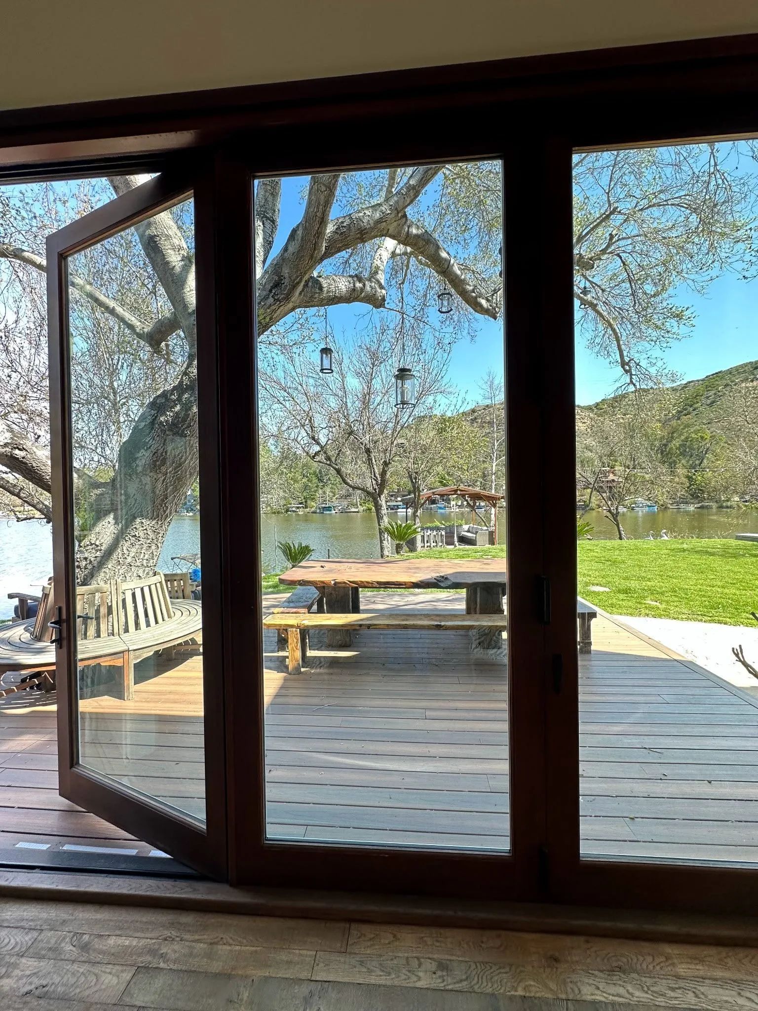 Open glass doors framing a sunny deck, large tree, and outdoor waterfront view.
