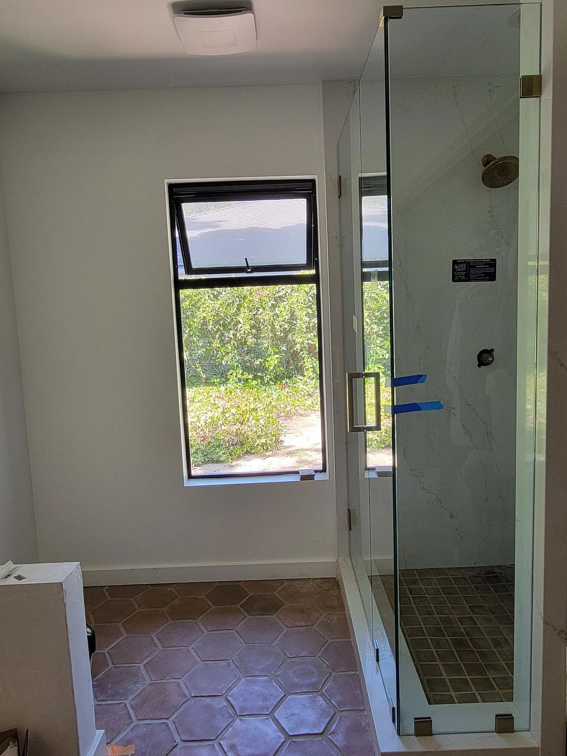 Small bathroom with glass shower, window, white walls, and brown tile floor.